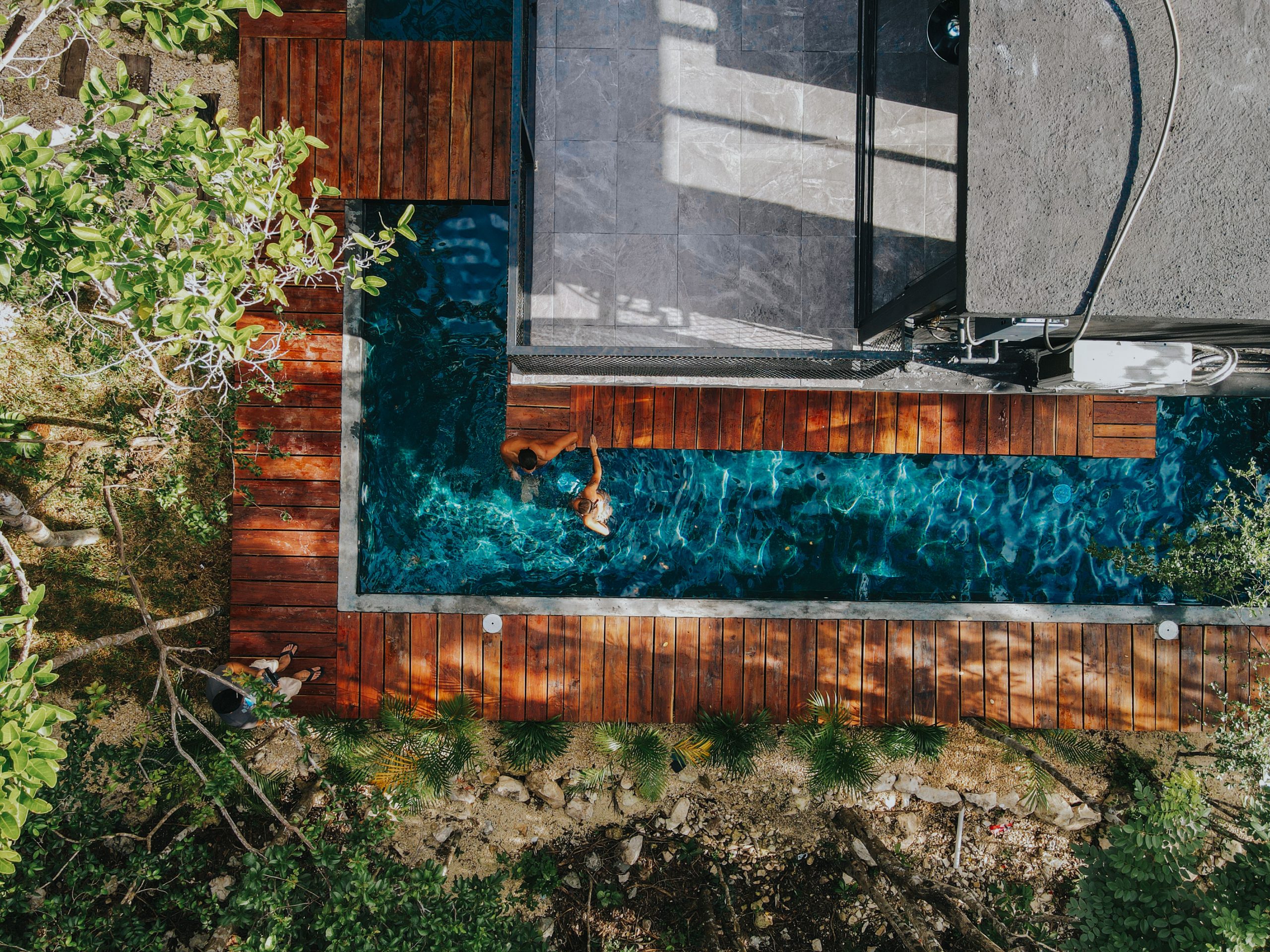 Aerial drone shot of a lap pool surrounded by modern wooden decking and lush green tropical foliage.