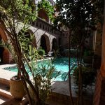 View of a turquoise clear water pool at the heart of a traditional Moroccan riad, surrounded by lush plants, mudbrick arches, and a brick patio under the summer sun.