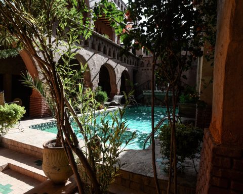 View of a turquoise clear water pool at the heart of a traditional Moroccan riad, surrounded by lush plants, mudbrick arches, and a brick patio under the summer sun.