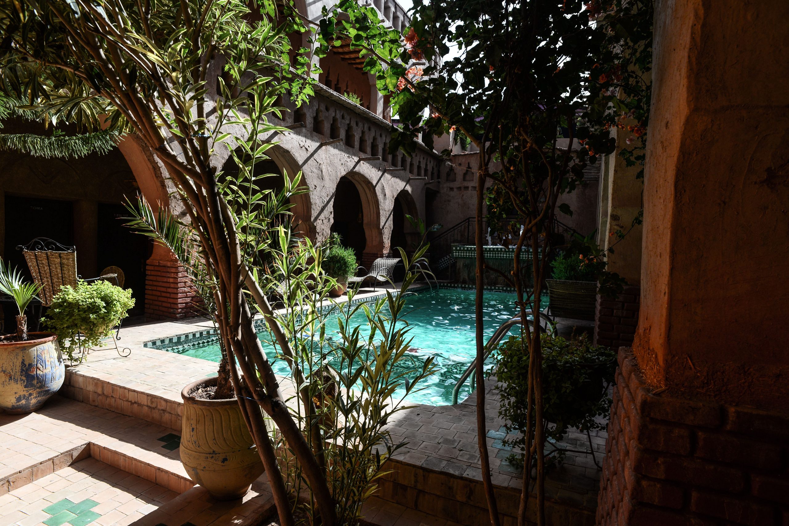 View of a turquoise clear water pool at the heart of a traditional Moroccan riad, surrounded by lush plants, mudbrick arches, and a brick patio under the summer sun.