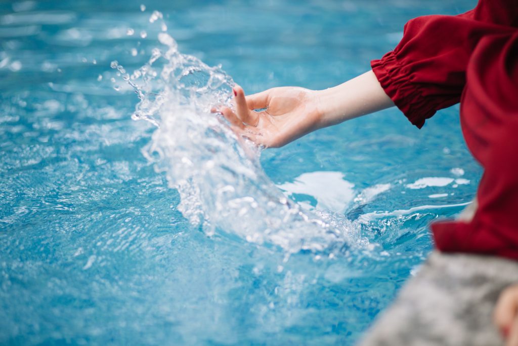Woman's hand dipping into a clear blue swimming pool, creating a wave of water splash.
