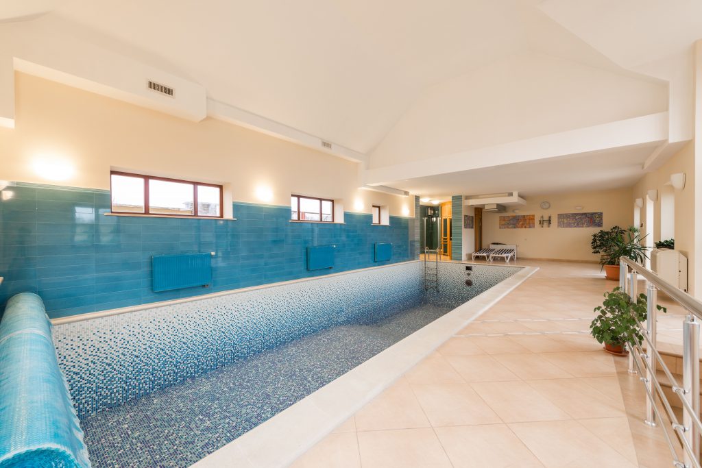 Wide shot of an empty indoor swimming pool with blue mosaic tiles and a thermal cover rolled up.
