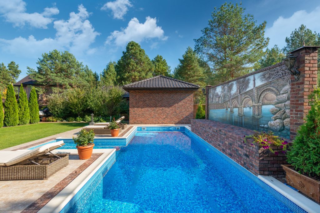 A rectangular blue tiled swimming pool with a brick wall, stone patio, lounge chairs, and manicured garden.