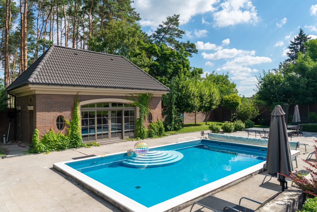 A private in-ground swimming pool with circular steps and a rainbow beach ball, positioned in front of a brick pool house with large windows.