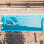 Overhead drone photograph looking straight down at a swimmer mid-stroke in a rectangular pool surrounded by grey stone paving.