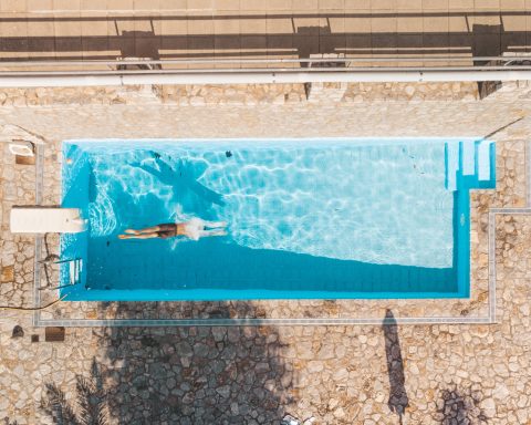 Overhead drone photograph looking straight down at a swimmer mid-stroke in a rectangular pool surrounded by grey stone paving.