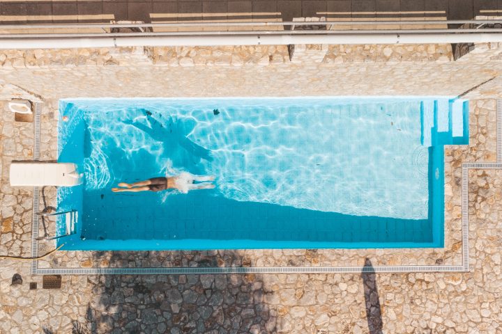 Overhead drone photograph looking straight down at a swimmer mid-stroke in a rectangular pool surrounded by grey stone paving.