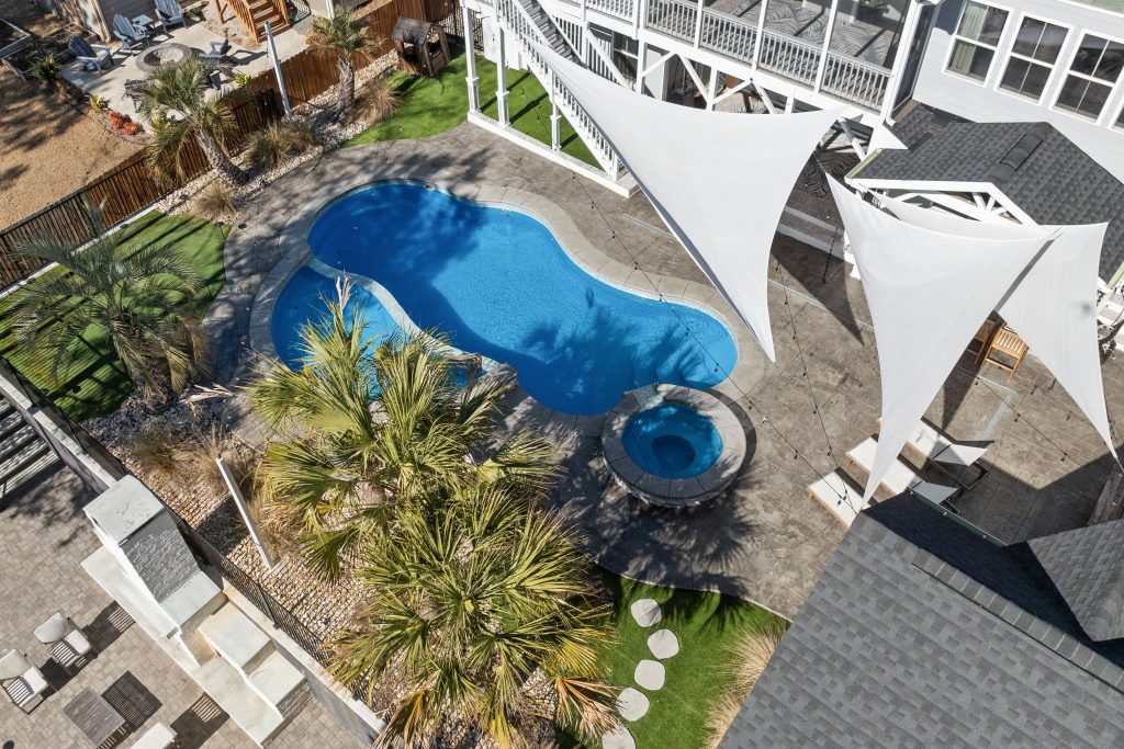 A residential backyard with a rectangular swimming pool, green lawn, and a large wooden pergola shading a deck with chairs, showing neighboring houses.