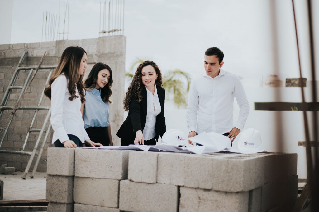 Four architects and engineers in professional attire discussing blueprints resting on cinder blocks at an active building construction site.