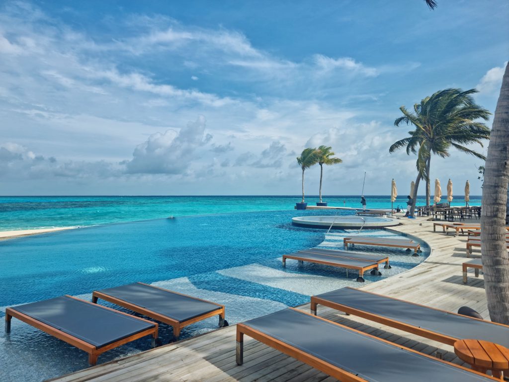 High-angle view of a large resort infinity pool with wooden deck sun loungers facing a turquoise ocean under a cloudy blue sky.