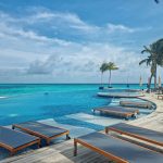 High-angle view of a large resort infinity pool with wooden deck sun loungers facing a turquoise ocean under a cloudy blue sky.