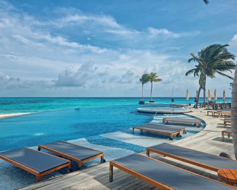 High-angle view of a large resort infinity pool with wooden deck sun loungers facing a turquoise ocean under a cloudy blue sky.