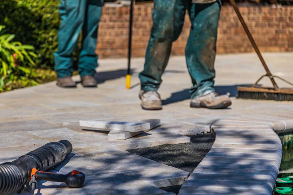 Close-up of two workers installing light-colored stone pavers for a pool coping, with tools visible.