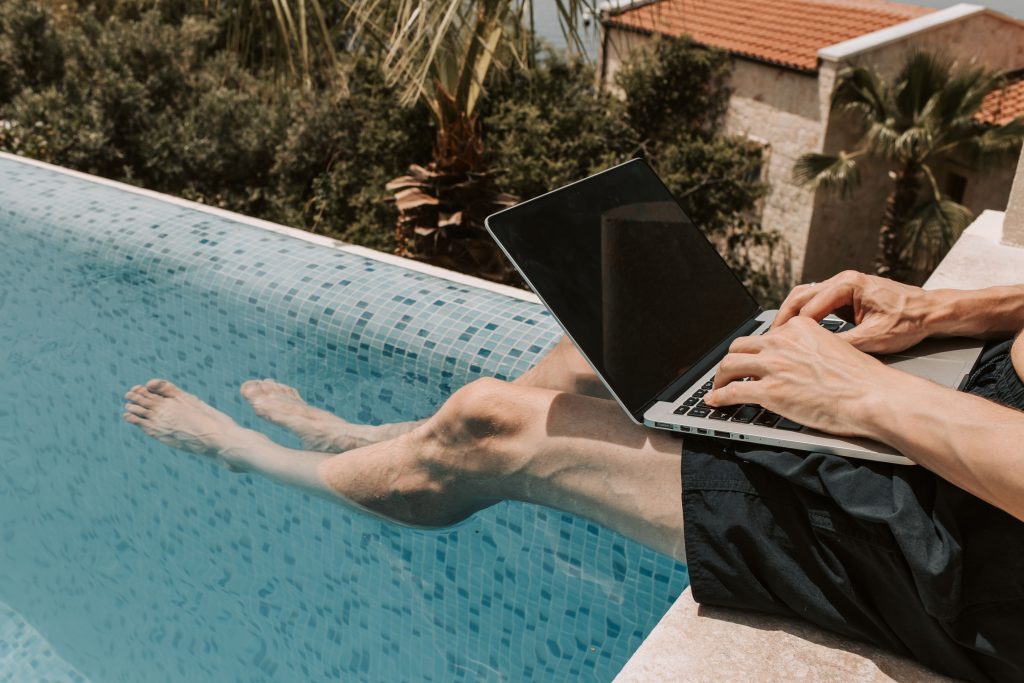 A traveler relaxing by the edge of a swimming pool with their legs in the water while using a laptop.