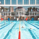 People swimming in lanes at a large outdoor public swimming pool with lane lines and a modern glass building in the background.