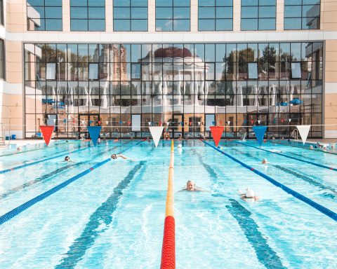People swimming in lanes at a large outdoor public swimming pool with lane lines and a modern glass building in the background.