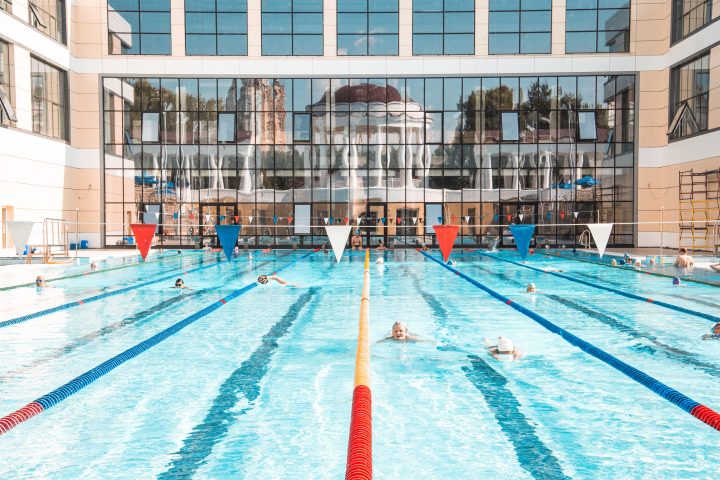 People swimming in lanes at a large outdoor public swimming pool with lane lines and a modern glass building in the background.