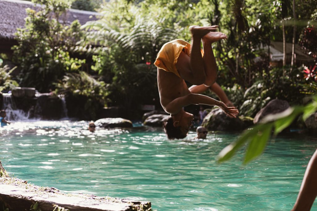 Person performing a backflip jump into a biological pool surrounded by rocks and trees.