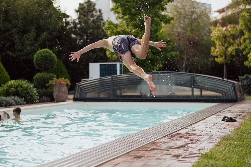 A young boy diving mid-air into a backyard swimming pool with a retractable cover.