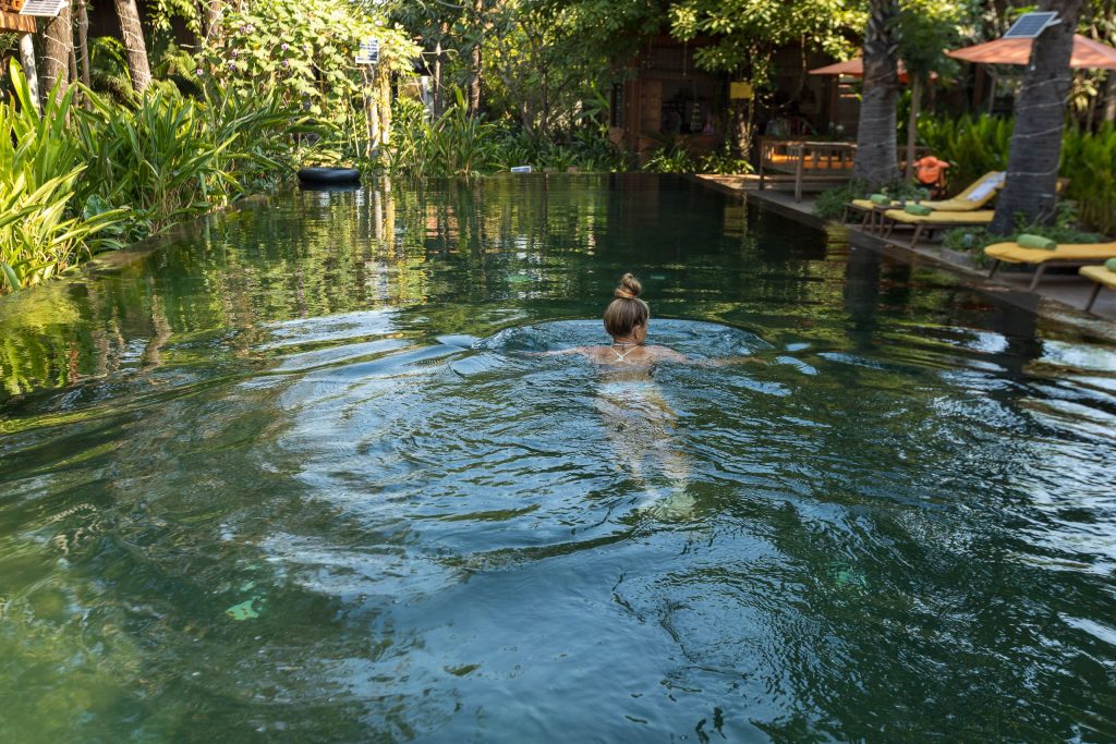 Woman swimming in dark green natural pool water surrounded by patio chairs and lush garden trees.