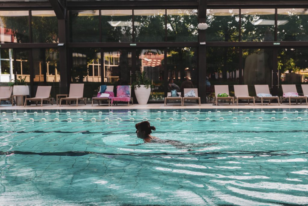 A woman swimming breaststroke in an outdoor public pool, with a row of empty lounge chairs and a large glass-fronted building with trees behind them.
