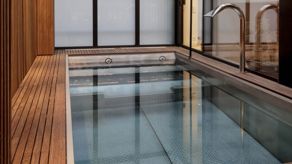 Indoor stainless steel pool with wooden deck and chrome fixtures by L'Esprit Piscine
