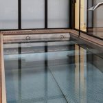 Indoor stainless steel pool with wooden deck and chrome fixtures by L'Esprit Piscine