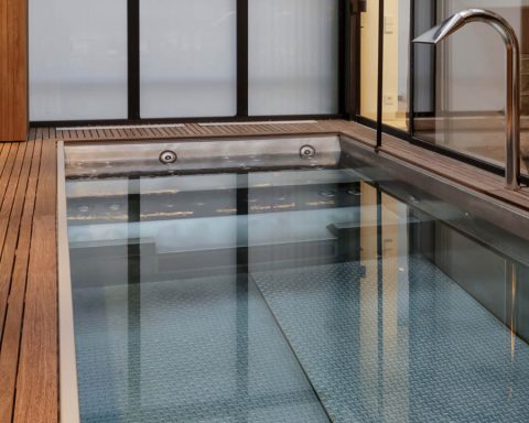 Indoor stainless steel pool with wooden deck and chrome fixtures by L'Esprit Piscine