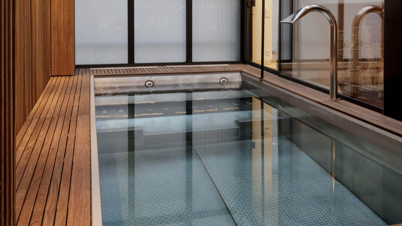 Indoor stainless steel pool with wooden deck and chrome fixtures by L'Esprit Piscine