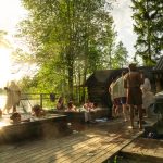 People relaxing in outdoor hot tubs and cooling down on a wooden dock by a lake at sunset.