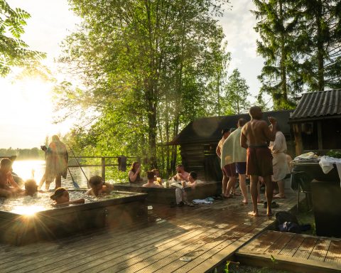 People relaxing in outdoor hot tubs and cooling down on a wooden dock by a lake at sunset.