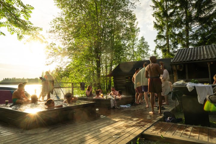 People relaxing in outdoor hot tubs and cooling down on a wooden dock by a lake at sunset.