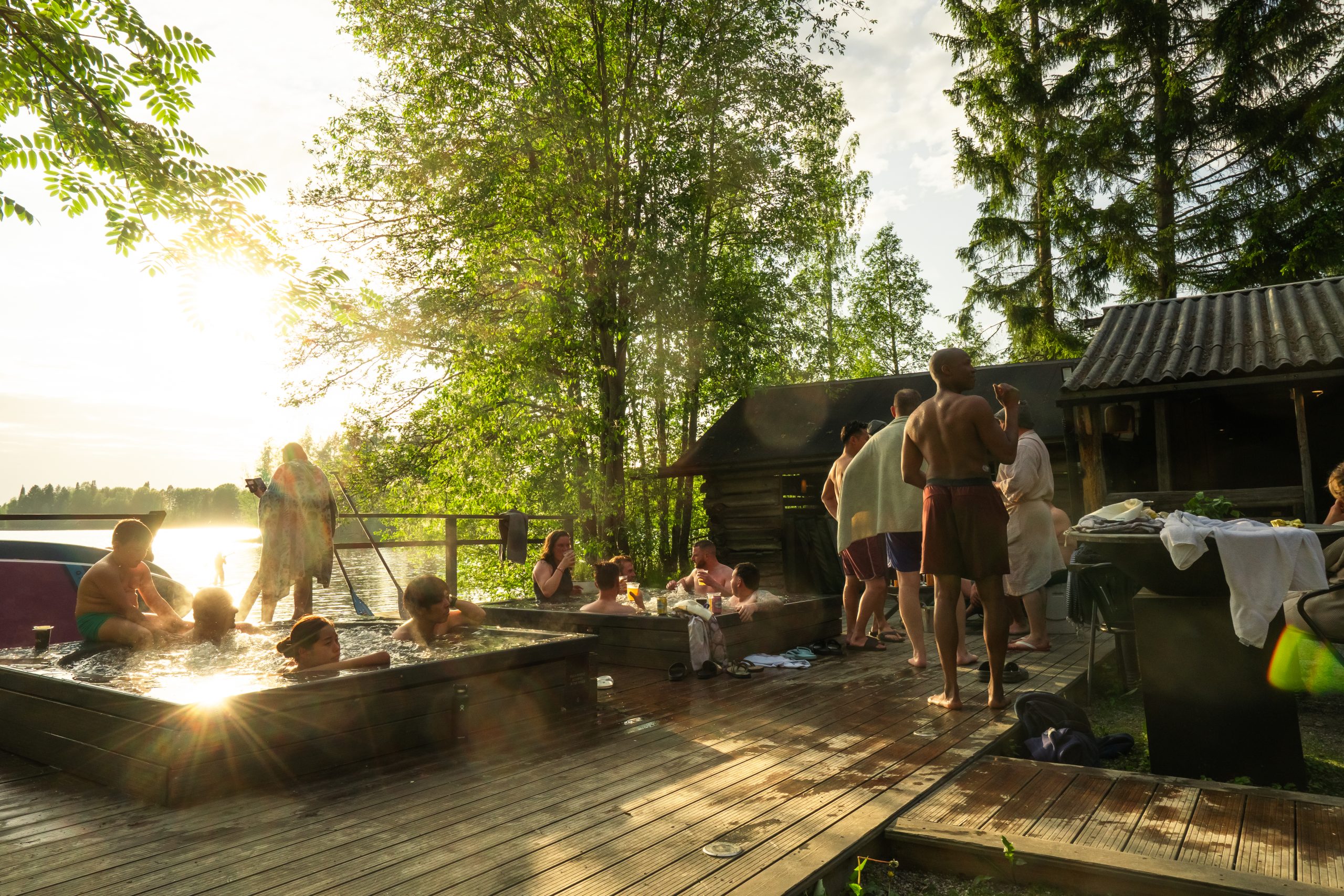 People relaxing in outdoor hot tubs and cooling down on a wooden dock by a lake at sunset.