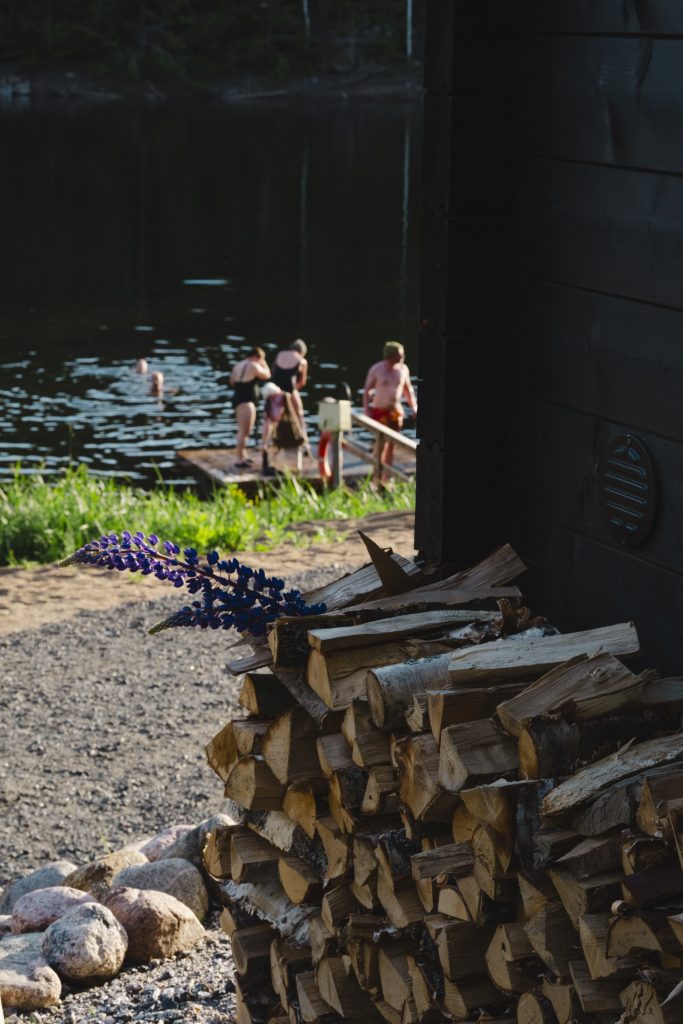 Stacked logs of firewood near a lake with people swimming after a sauna session.