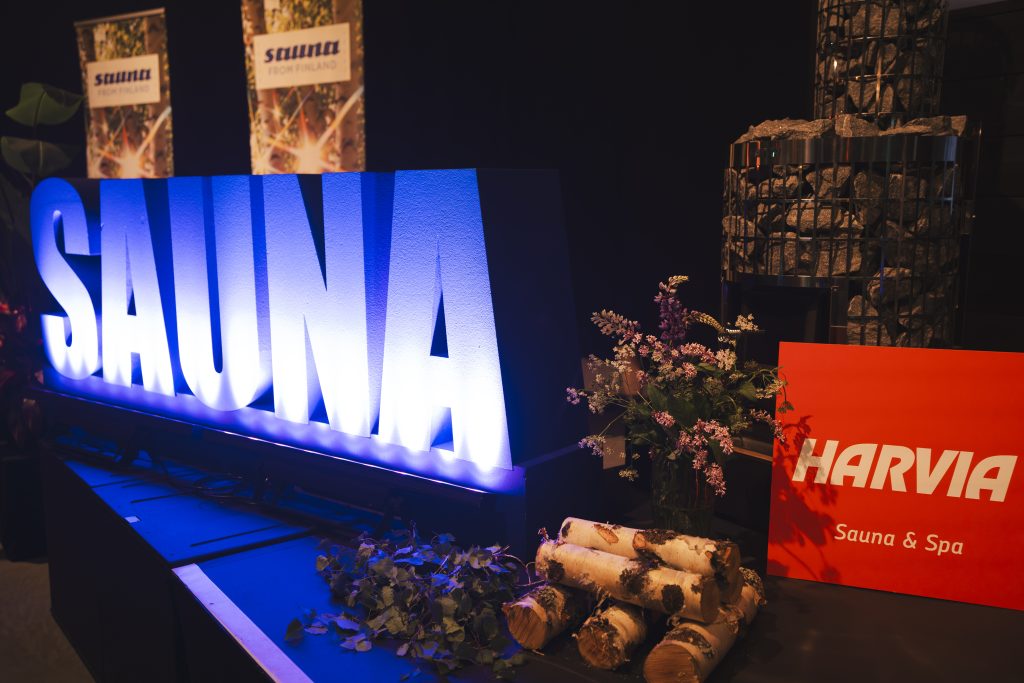 Blue illuminated SAUNA letters next to birch logs, sauna whisks, and a Harvia sign at an exhibition.