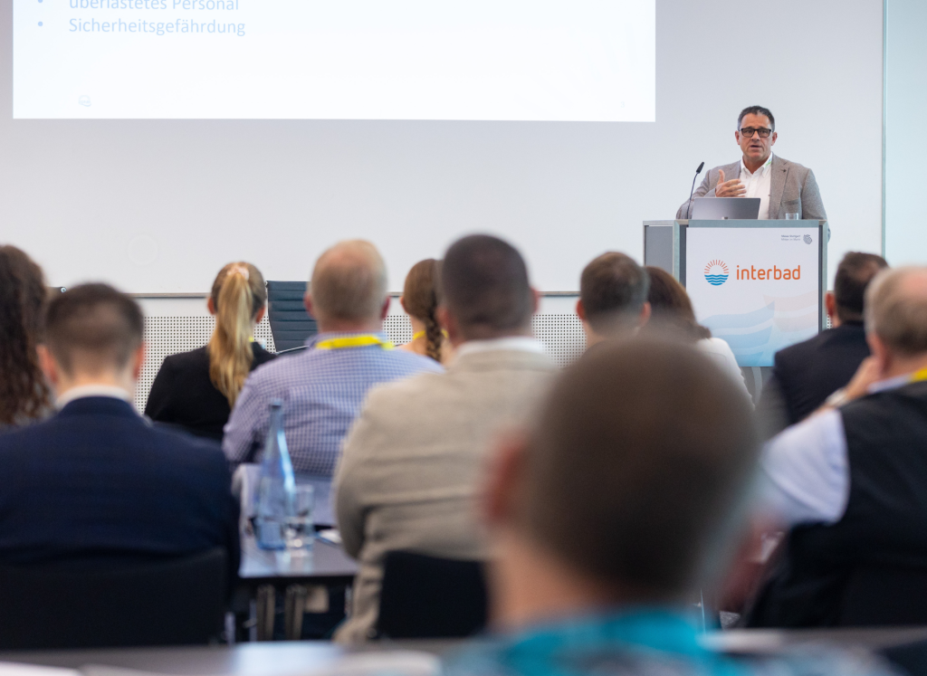 A speaker presenting at a lectern with the interbad logo during a professional conference session, viewed from behind the audience.
