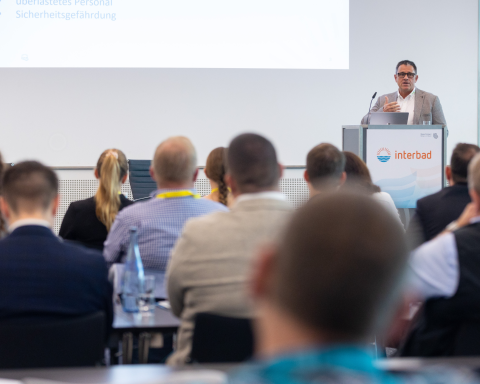 A speaker presenting at a lectern with the interbad logo during a professional conference session, viewed from behind the audience.