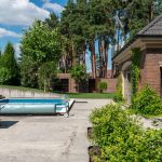 A rectangular residential swimming pool in a backyard patio with a blue solar bubble cover rolled up on a manual roller.