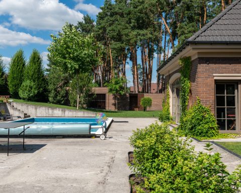 A rectangular residential swimming pool in a backyard patio with a blue solar bubble cover rolled up on a manual roller.