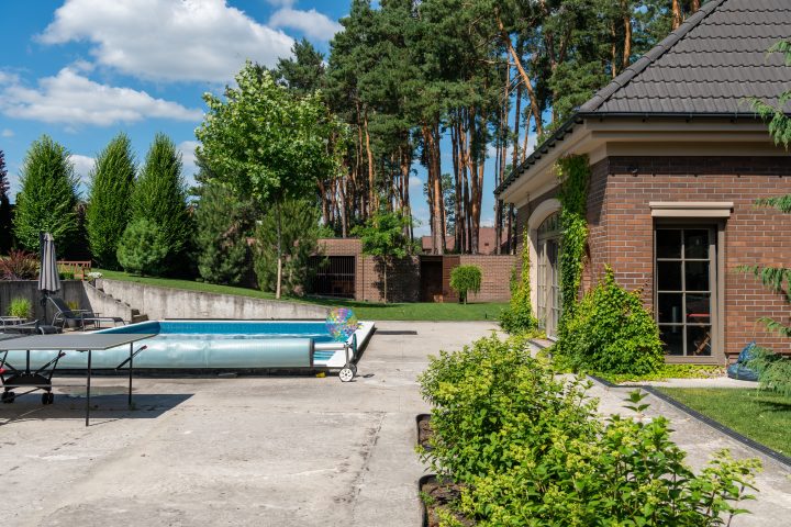A rectangular residential swimming pool in a backyard patio with a blue solar bubble cover rolled up on a manual roller.