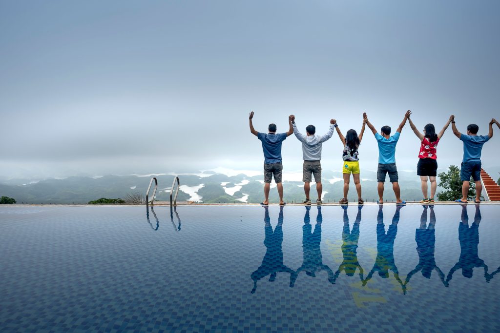 Six people standing side-by-side with raised arms at the edge of an infinity pool overlooking a foggy landscape.