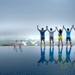 Six people standing side-by-side with raised arms at the edge of an infinity pool overlooking a foggy landscape.