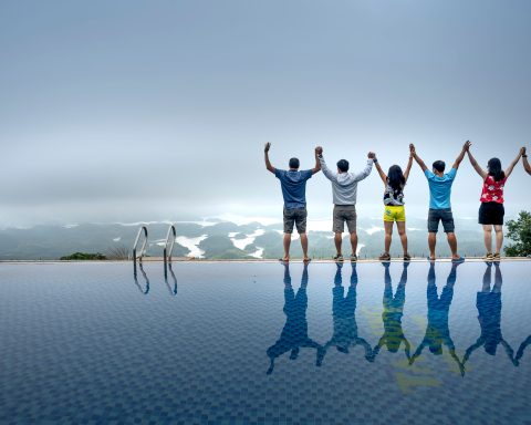 Six people standing side-by-side with raised arms at the edge of an infinity pool overlooking a foggy landscape.