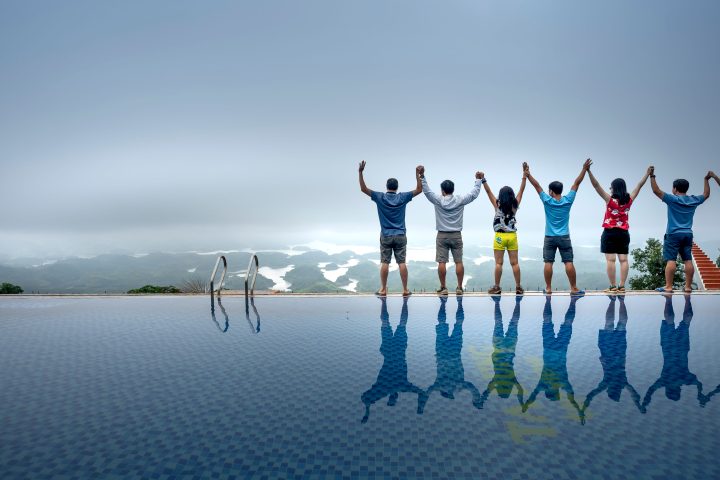 Sechs Personen stehen Seite an Seite mit erhobenen Armen am Rand eines Infinity-Pools und blicken auf eine neblige Landschaft.