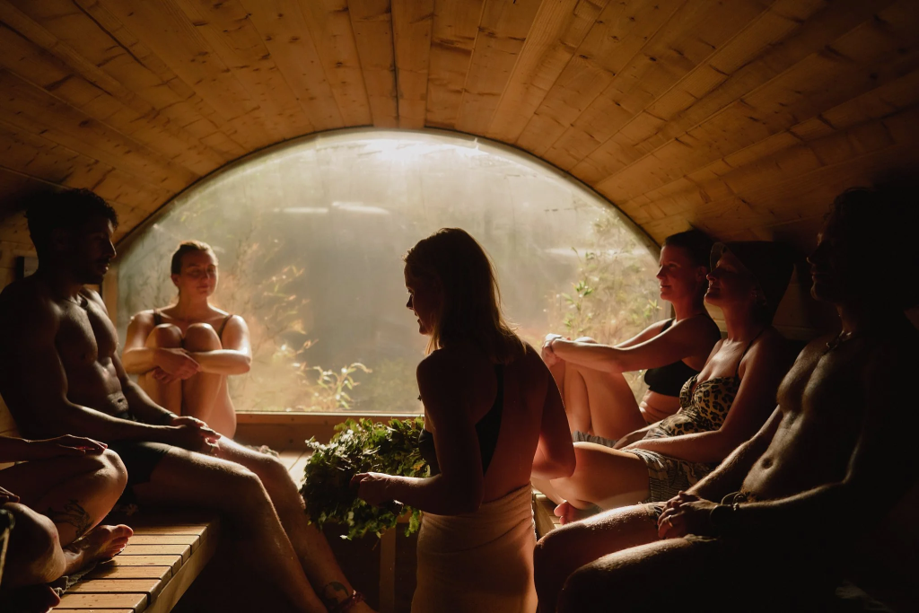 Group of bathers seated inside a wooden barrel sauna with a large circular window facing nature