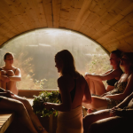 Group of bathers seated inside a wooden barrel sauna with a large circular window facing nature