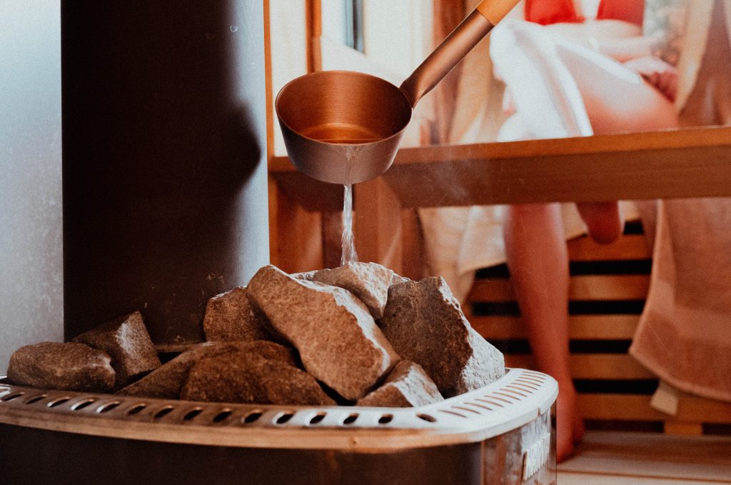 Copper ladle pouring water onto hot sauna stones to generate steam inside a Finnish sauna