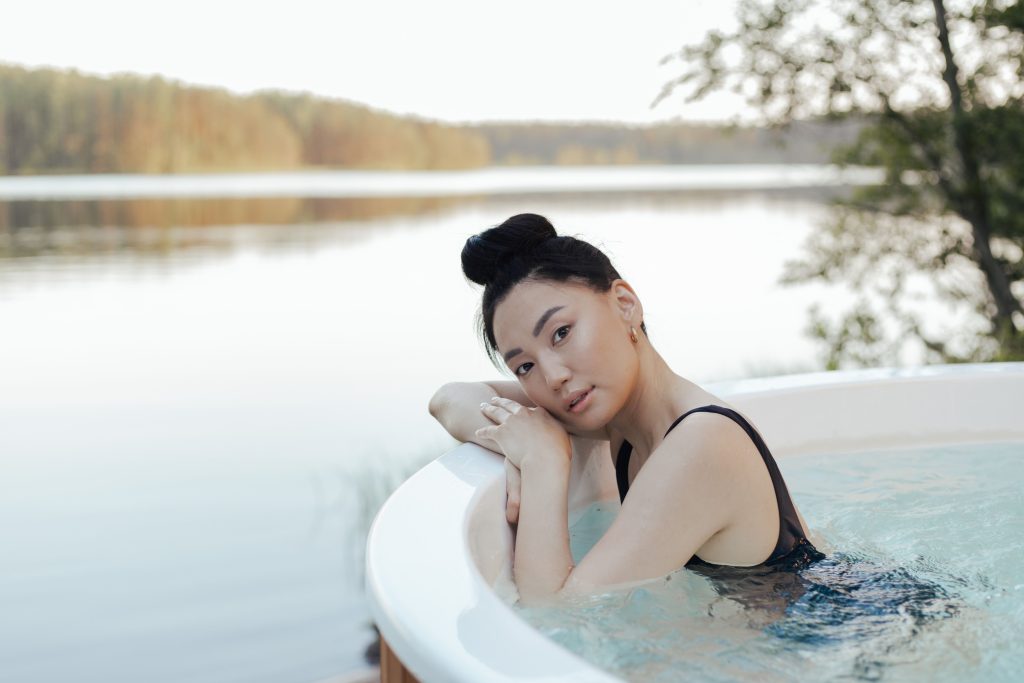 A woman smiling while relaxing inside a round wooden hot tub facing a serene natural lake landscape