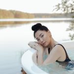 A woman smiling while relaxing inside a round wooden hot tub facing a serene natural lake landscape