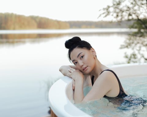 A woman smiling while relaxing inside a round wooden hot tub facing a serene natural lake landscape